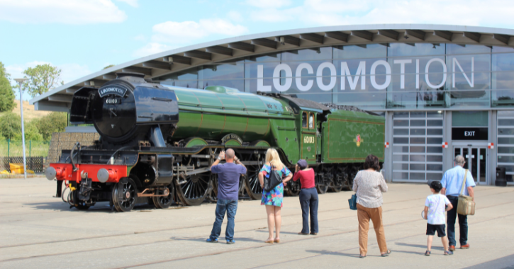 The Flying Scotsman on display outside Locomotion museum building surrounded by people taking photos and admiring the historic steam engine.